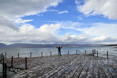 Rear view of man standing on pier over lake against cloudy sky