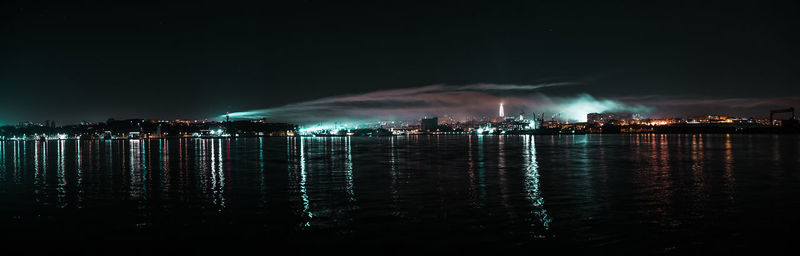 Illuminated city by sea against sky at night