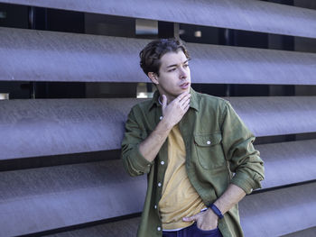 Young man in casual clothes standing outside with hand in pocket touching chin