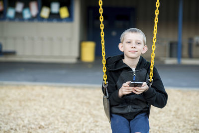 Portrait of boy using mobile phone while swinging at park