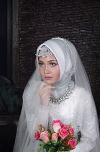 Bride wearing traditional clothing holding bouquet against wall