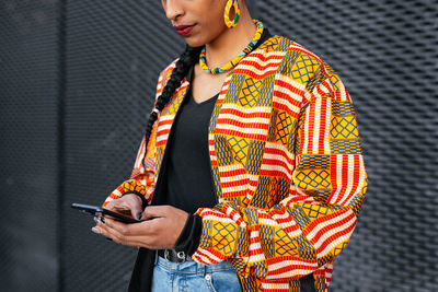 Cropped unrecognizable trendy young ethnic female in colorful clothes browsing on smartphone against gray wall on city street