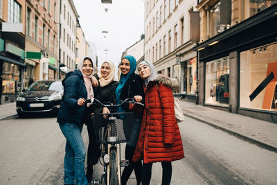 Portrait of smiling multi-ethnic muslim female friends standing with bicycle on street in city