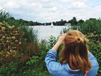 Rear view of woman photographing sailboat in lake against cloudy sky