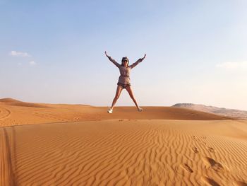 Cheerful woman with arms outstretched jumping at desert against sky