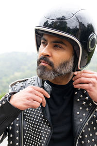 Self assured adult bearded male biker in stylish leather jacket adjusting protective helmet and looking away in nature