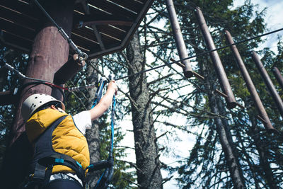 Below view of kid attaching safety harness on zip line while standing on treetop in adventure park.