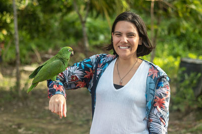 Woman poses with a smile as she carries her arm to a macaw in a camp