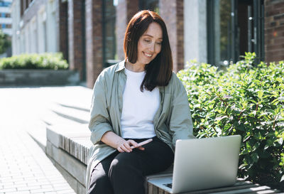 Adult  woman forty years in stylish shirt working on laptop at public place on bench at city street