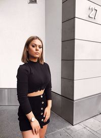 Portrait of young woman standing on footpath against wall