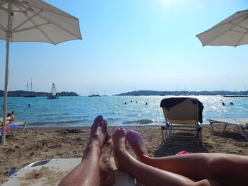 Low section of couple relaxing at beach against clear sky