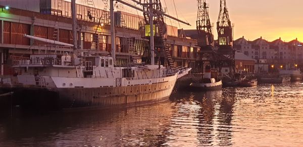 Sailboats moored on river in city against sky during sunset