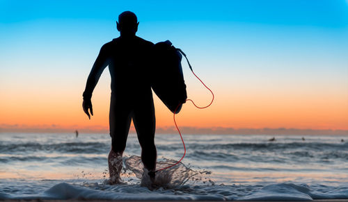 Rear view of man carrying equipment while walking in sea against sky during sunset