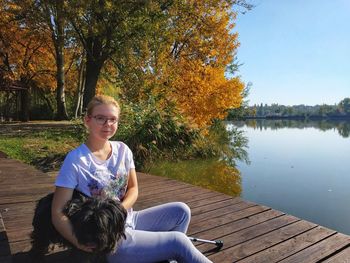 Portrait of woman with dog sitting on pier over lake against trees