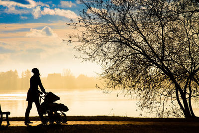 Silhouette woman with baby stroller walking on footpath against sky during sunset