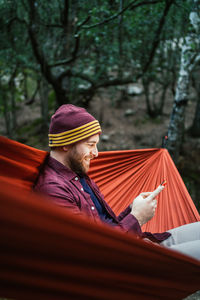 Young male uses his smartphone resting on a hammock in the woods