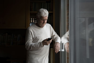 Senior mam using phone while standing by window at home