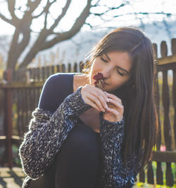 Close-up of woman smelling flower while sitting against fence