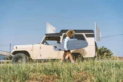 Young woman carrying surfboard by old off-road vehicle on sunny day