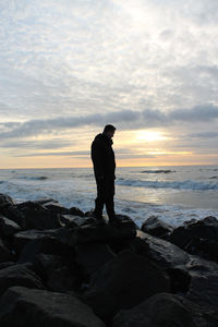 Thoughtful man standing on rocky shore against cloudy sky during sunset