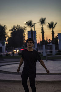 Portrait of young man standing on footpath in city at sunset