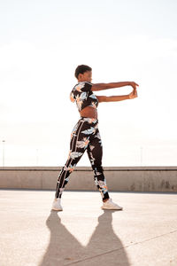 From below focused african american adult woman in flowered sportswear stretching hand muscles while standing alone and warming up before training among urban environment in sunny day