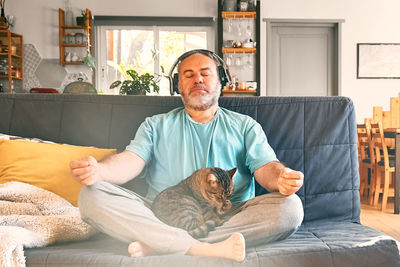 Middle-aged overweight man in wireless headphones relaxing at home with cat and guided meditation.