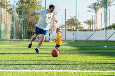 Joyful father with daughter playing football on green sports field.