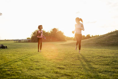 Happy girl jogging with mother on grass at park against sky during sunset