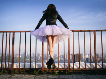 A ballerina in a tutu  jumps holding on to the fence at sunset admiring the panorama of the city