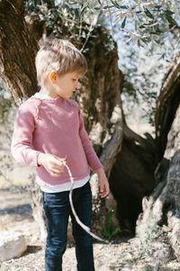Full length of cute boy holding twig standing against tree