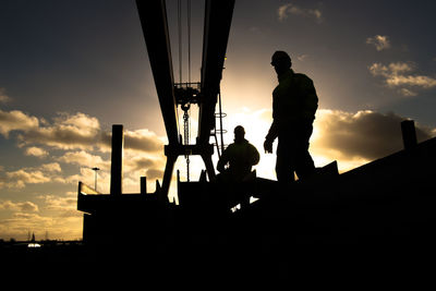 Silhouette workers by metallic structure against cloudy sky during sunset