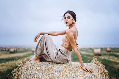 Portrait of young woman sitting on hay bale at farm