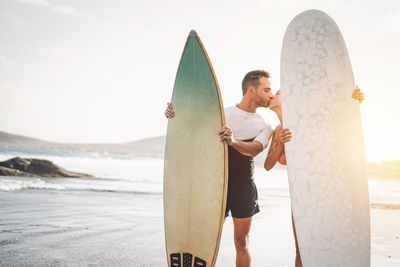 Couple kissing while standing at beach with surfboards