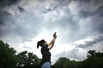 Low angle view of woman aiming with gun towards stormy clouds