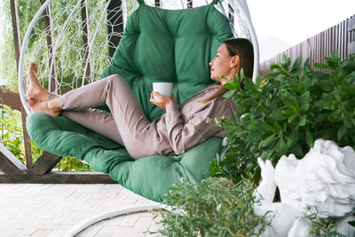 Real woman drinking tea or coffee outdoor. full length mature woman sitting in a hammock chair 