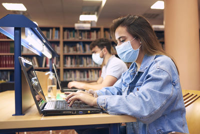 A girl and a boy wearing masks study with laptops in the library