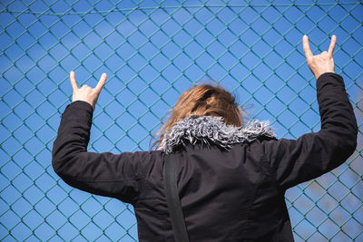 Rear view of woman showing horn sign by chainlink fence