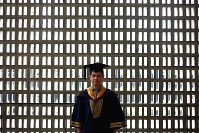 Portrait of man in graduation gown standing against window