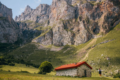 Woman standing in majestic landscape of picos de europa mountain range near small stone cabin located on green meadow on sunny day in asturias