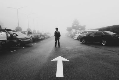 Rear view of man standing by arrow symbol amidst cars during foggy weather