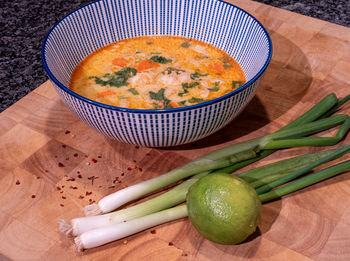 Asian coconut soup with red curry paste in a blue bowl on a wooden block with spring onions, lime.