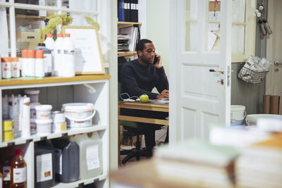 Confident young male owner talking on smart phone while sitting at desk in furniture store