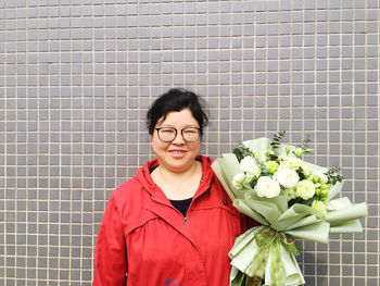 Portrait of smiling woman holding bouquet standing against wall