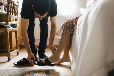 Young male healthcare worker helping senior woman to wear sandal in bedroom