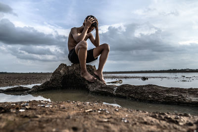 Sad boy sitting on rock by sea against cloudy sky