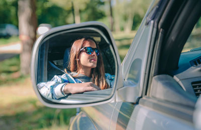Woman wearing sunglasses reflecting on side-view mirror in forest
