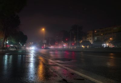 Wet road by illuminated city against sky at night