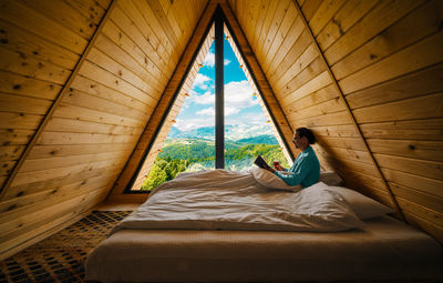 Man reading book and drinking wine in bed in a-frame wooden cabin at mountain