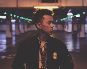 Young man looking away while standing against illuminated lights at night